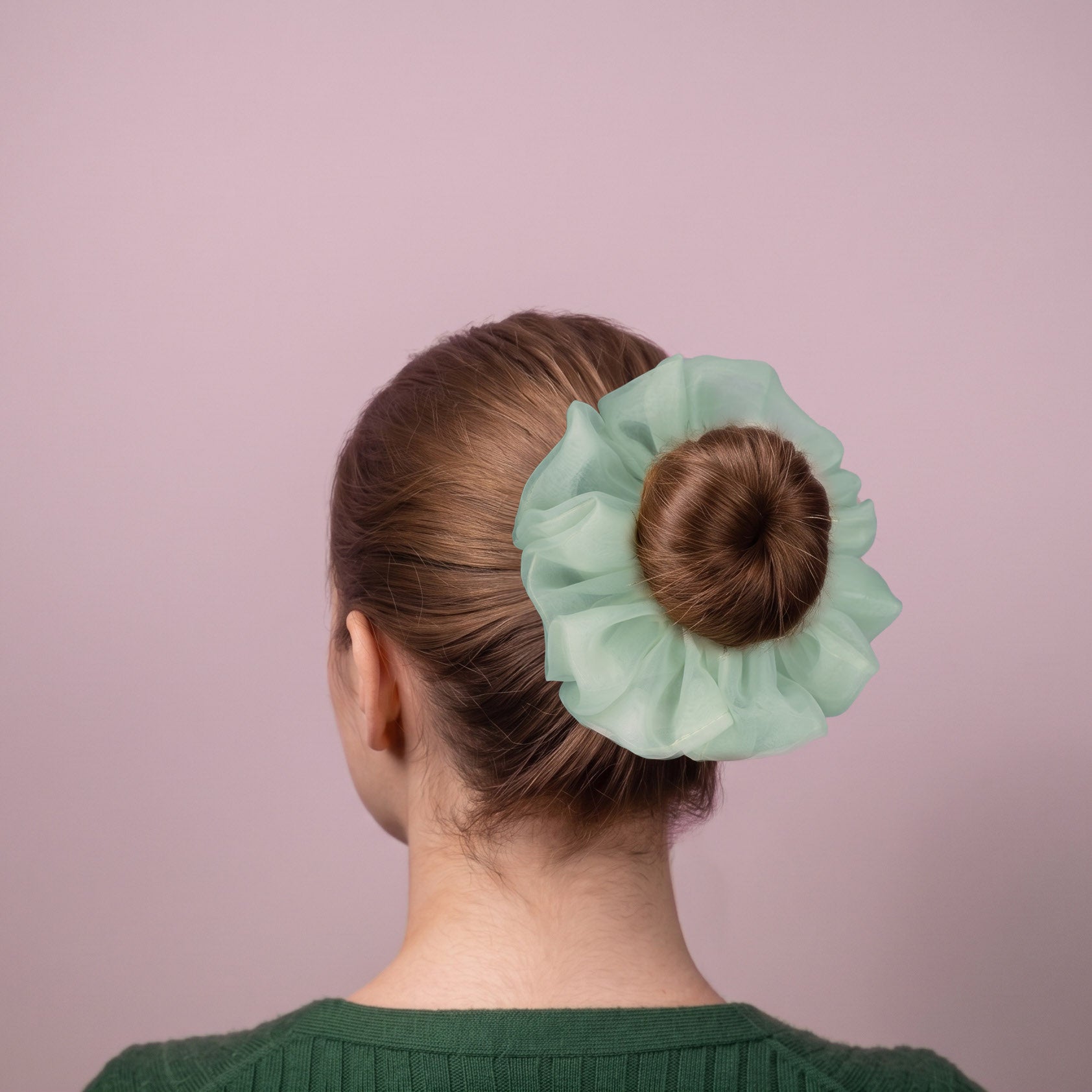 Person with a bun held by a light green organza scrunchie against a pink background